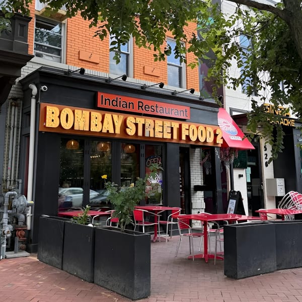 Exterior of Bombay Street Foods, an Indian restaurant, with red outdoor tables and black planters.
