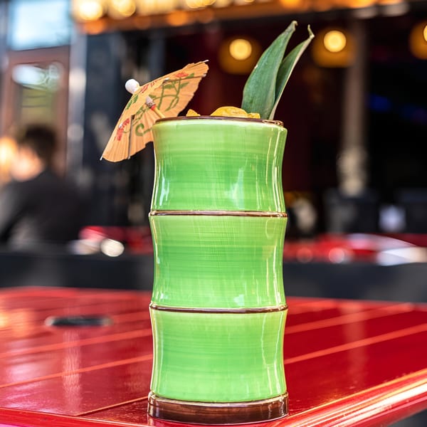 A vibrant green bamboo-shaped cocktail glass with a paper umbrella and pineapple leaves on a red table.