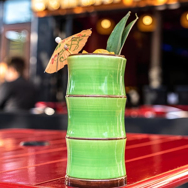 A green bamboo-shaped cocktail glass with a drink, pineapple leaves, and a small umbrella on a red table.