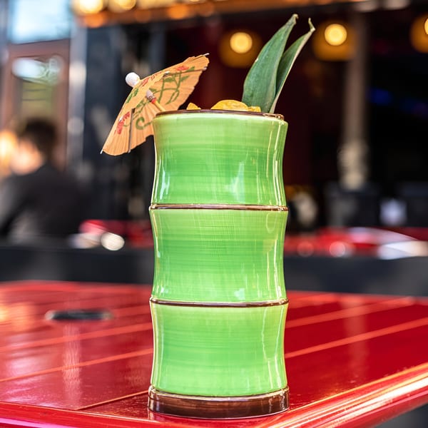 A vibrant green bamboo-shaped cocktail glass with a paper umbrella and pineapple leaves on a red table.