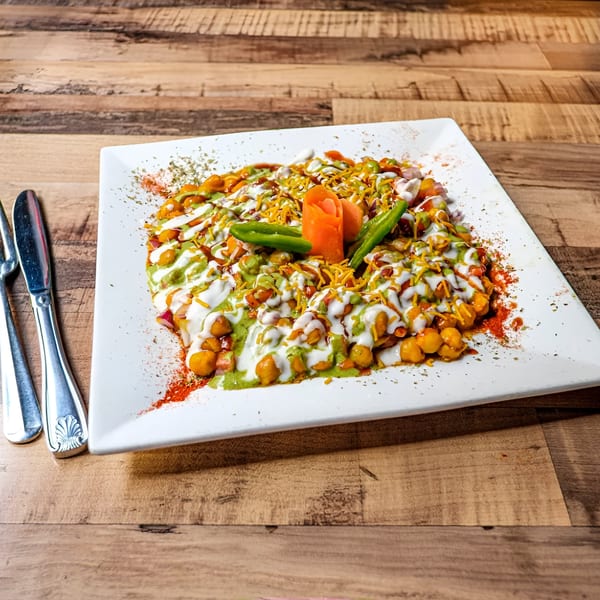 A Bombay Street Foods' Indian chaat dish, topped with sauces, chickpeas, and spices on a white plate.
