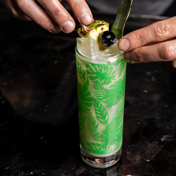 Hands garnishing a Bombay Street Foods cocktail with pineapple and a cherry in a green leaf-patterned glass.