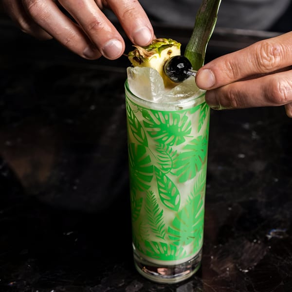 Hands garnishing a tall, green-patterned cocktail with pineapple, a cherry, and a leaf.