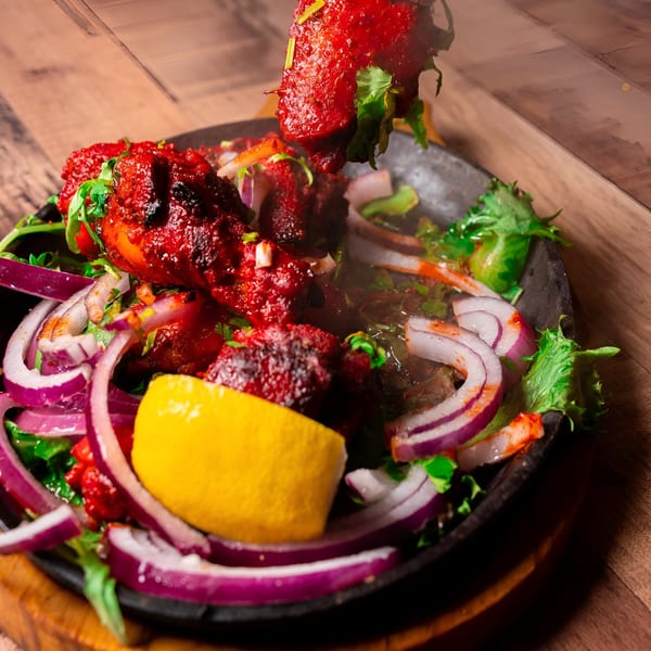 Sizzling red Indian tandoori chicken with onions, cilantro, and a lemon wedge on a dark platter.