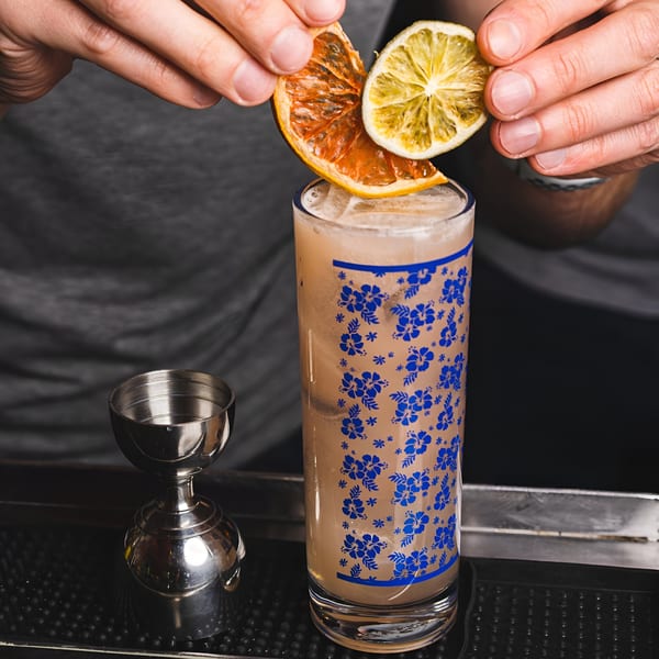 Hands garnishing a tall cocktail with dried orange and lime slices at Bombay Street Foods.