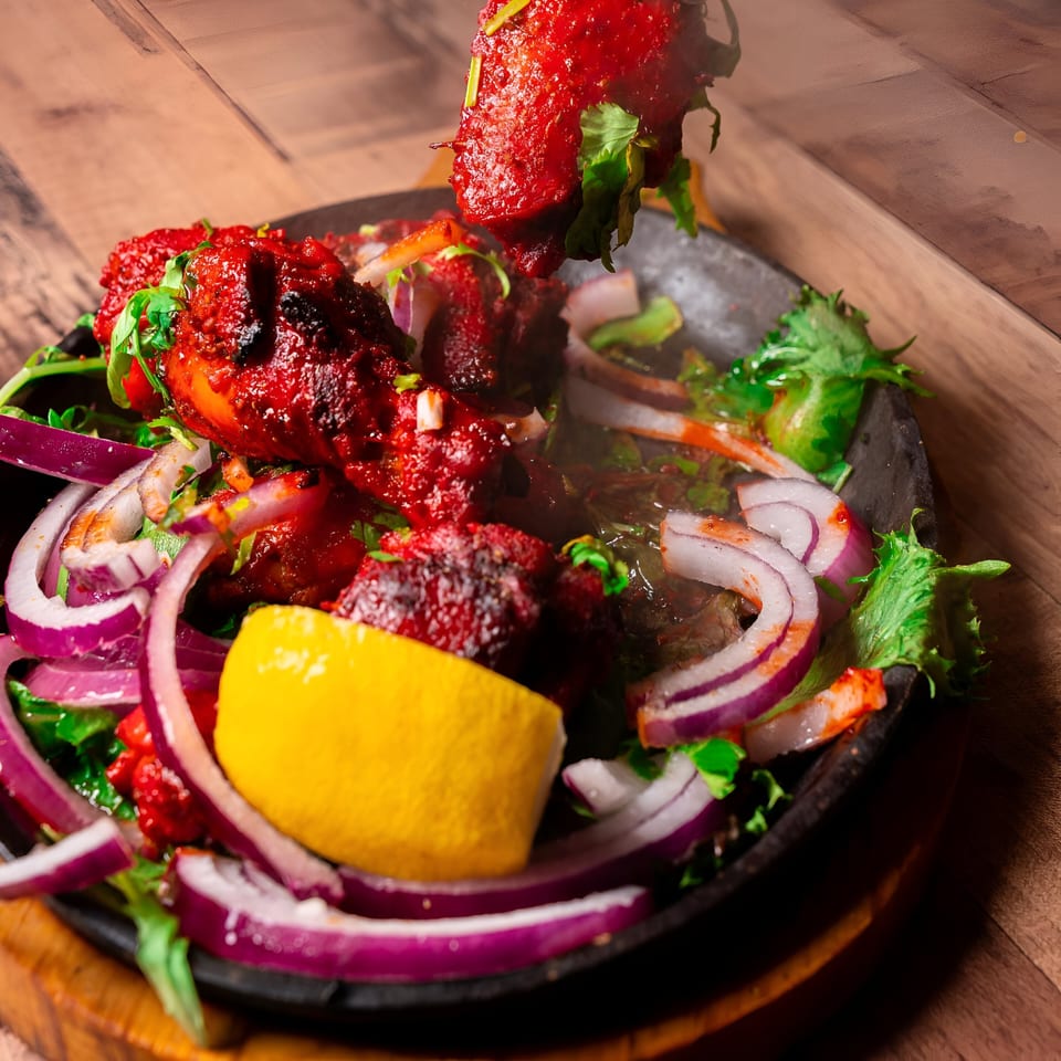 Sizzling red Indian chicken tikka with onions, cilantro, and lemon in a black skillet.