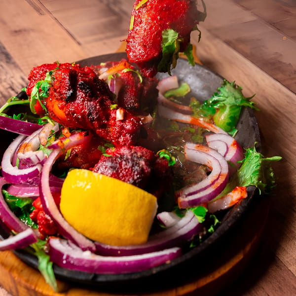Sizzling Indian chicken tikka with red onions, cilantro, and lemon in a black skillet.