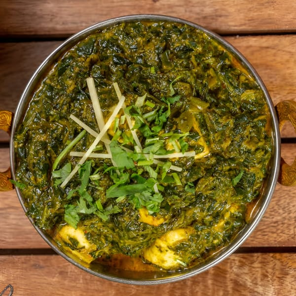 A metal bowl of vibrant green Palak Paneer, garnished with fresh cilantro and ginger, on a wooden table.